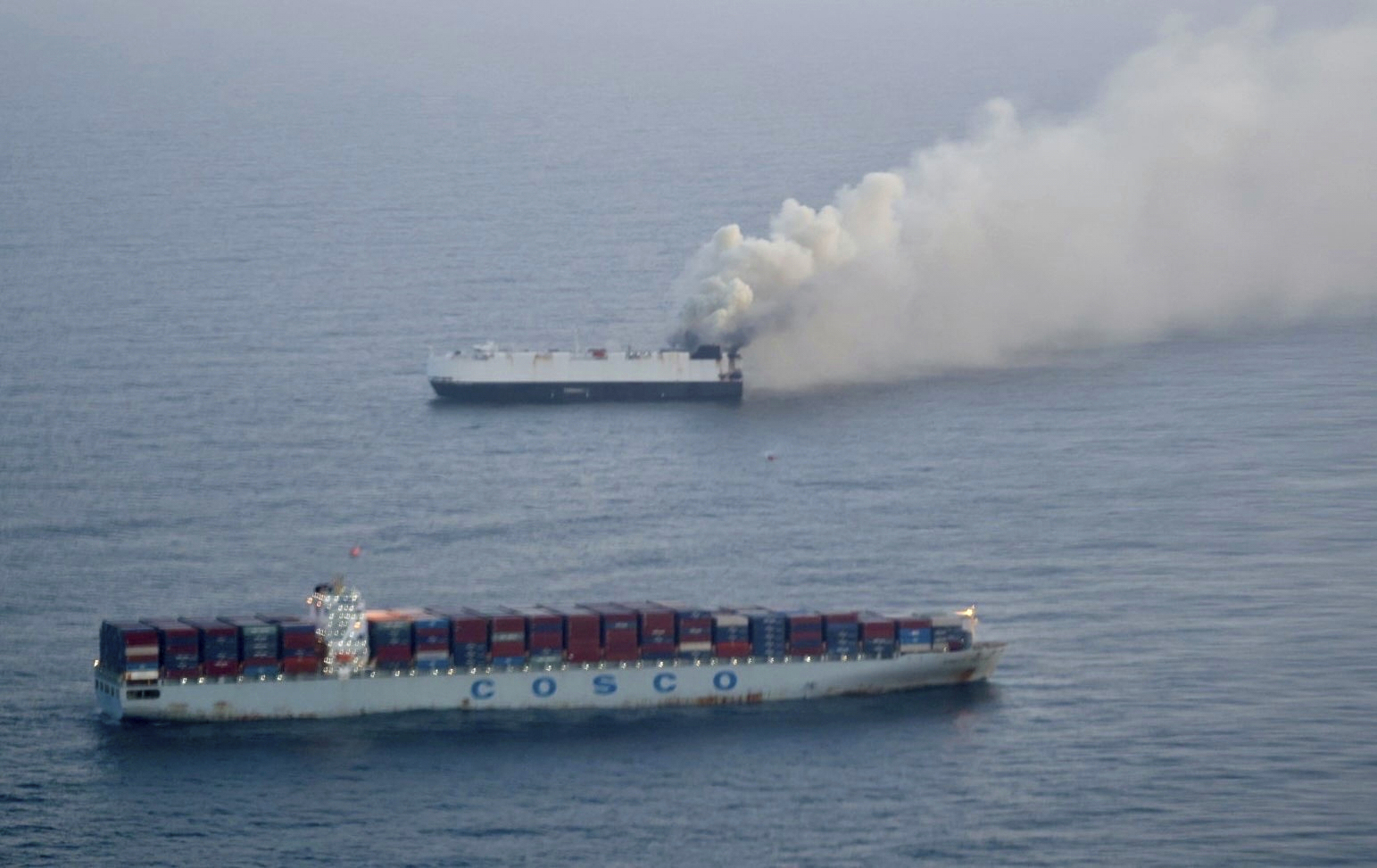 Two cargo ships at sea, one emitting thick smoke against a gray maritime horizon