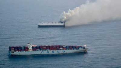Two cargo ships at sea, one emitting thick smoke against a gray maritime horizon