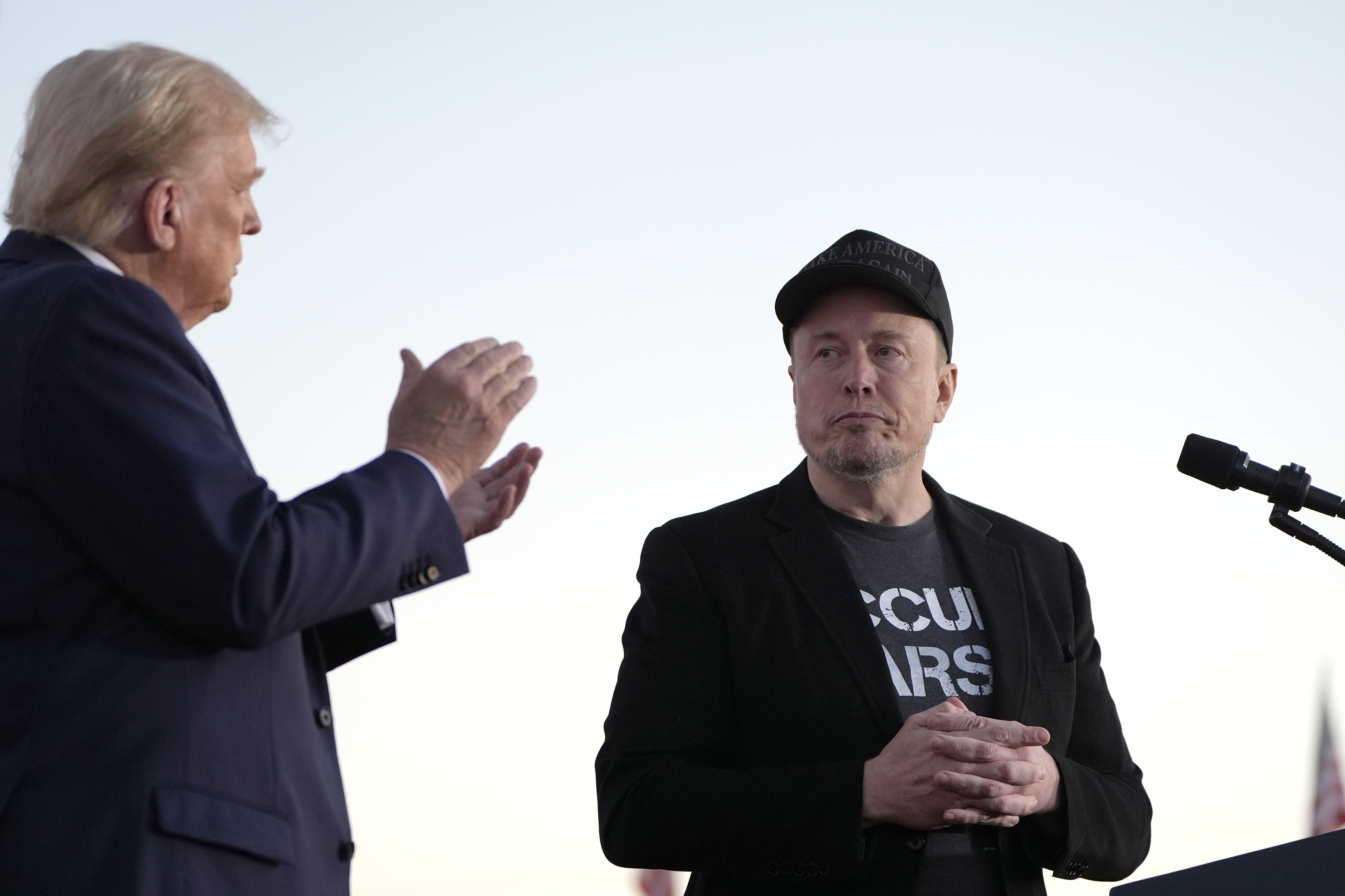 Political figures at a press conference, one applauding while the other stands stoically