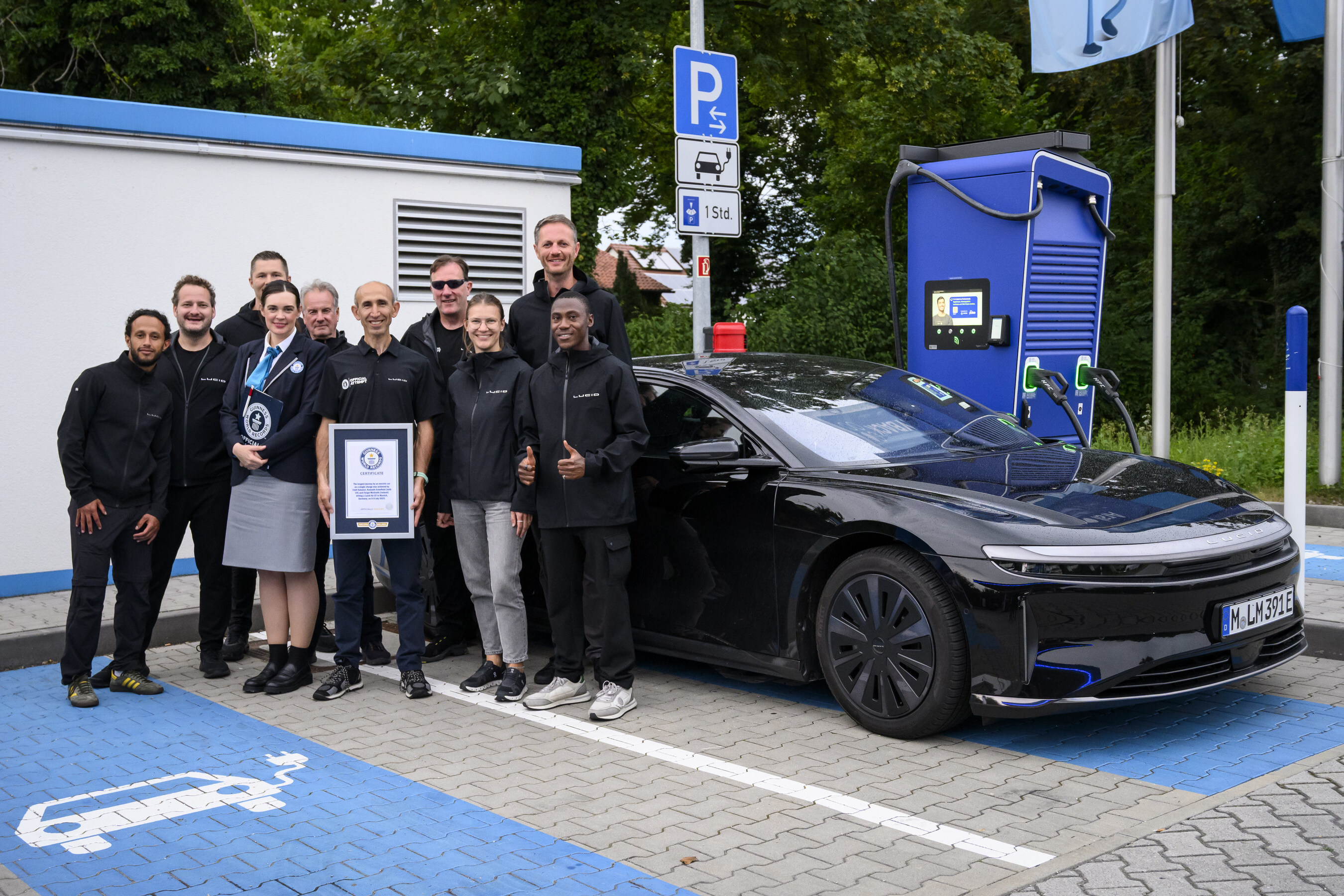 Group of people holding award certificates standing next to black electric vehicle at charging station with blue parking space markings