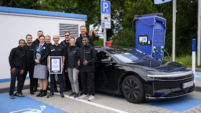 Group of people holding award certificates standing next to black electric vehicle at charging station with blue parking space markings