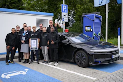 Group of people holding award certificates standing next to black electric vehicle at charging station with blue parking space markings