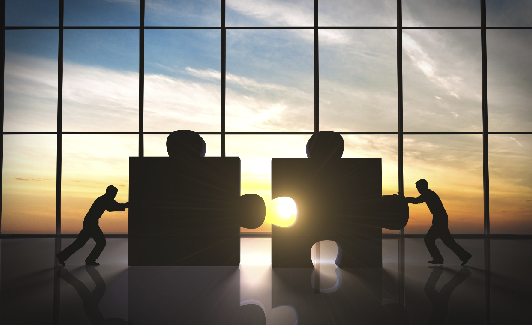 Silhouettes pushing puzzle pieces together at sunset, symbolizing teamwork and collaboration