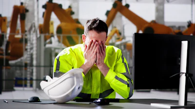 Worker in safety vest overwhelmed at desk with hard hat, industrial robots in background