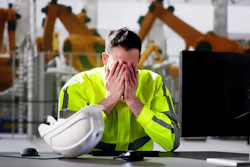 Worker in safety vest overwhelmed at desk with hard hat, industrial robots in background