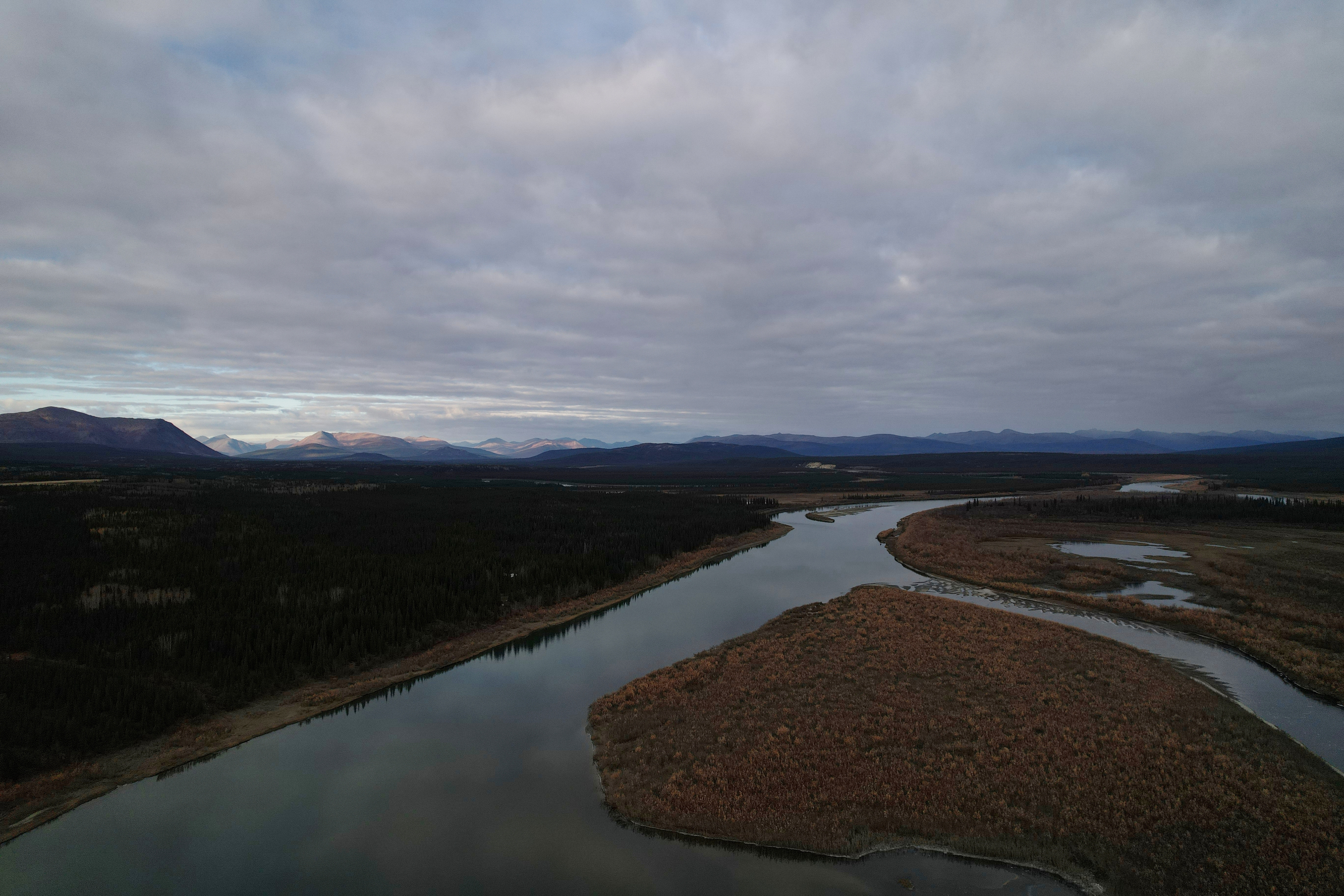 Winding river through forested valley with distant mountain ranges under cloudy sky
