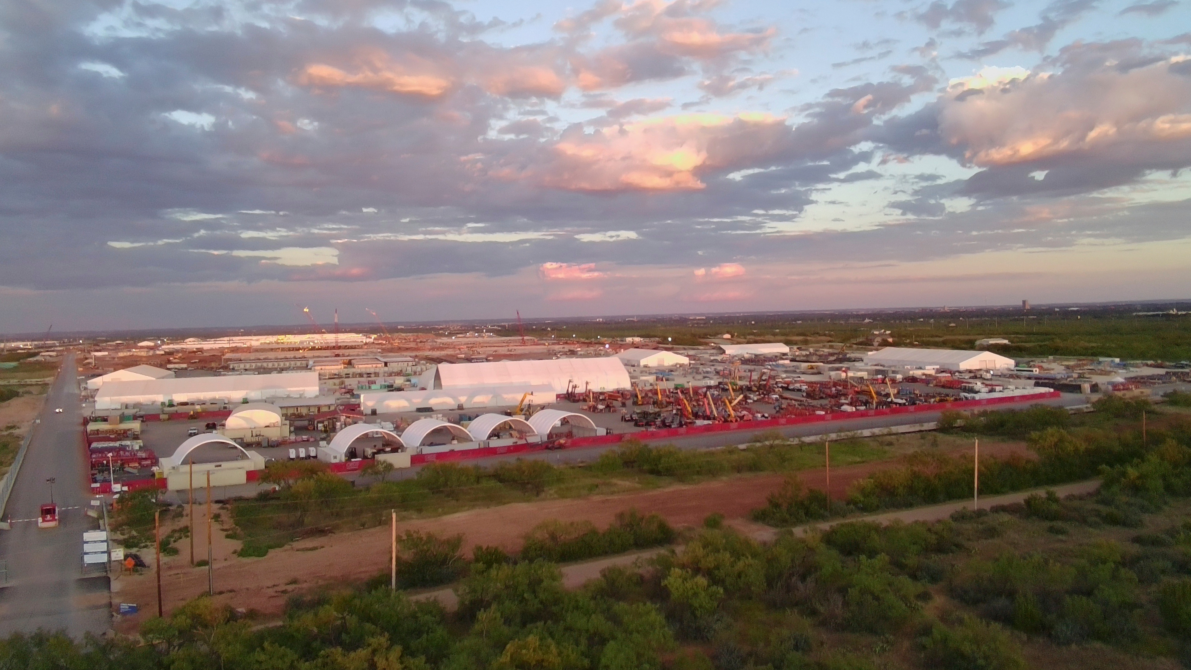Sunset over industrial complex with white arched buildings and red perimeter fence