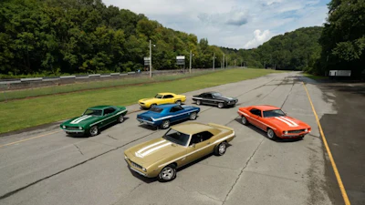 Six vintage muscle cars parked on an asphalt road with green forest background