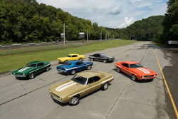 Six vintage muscle cars parked on an asphalt road with green forest background
