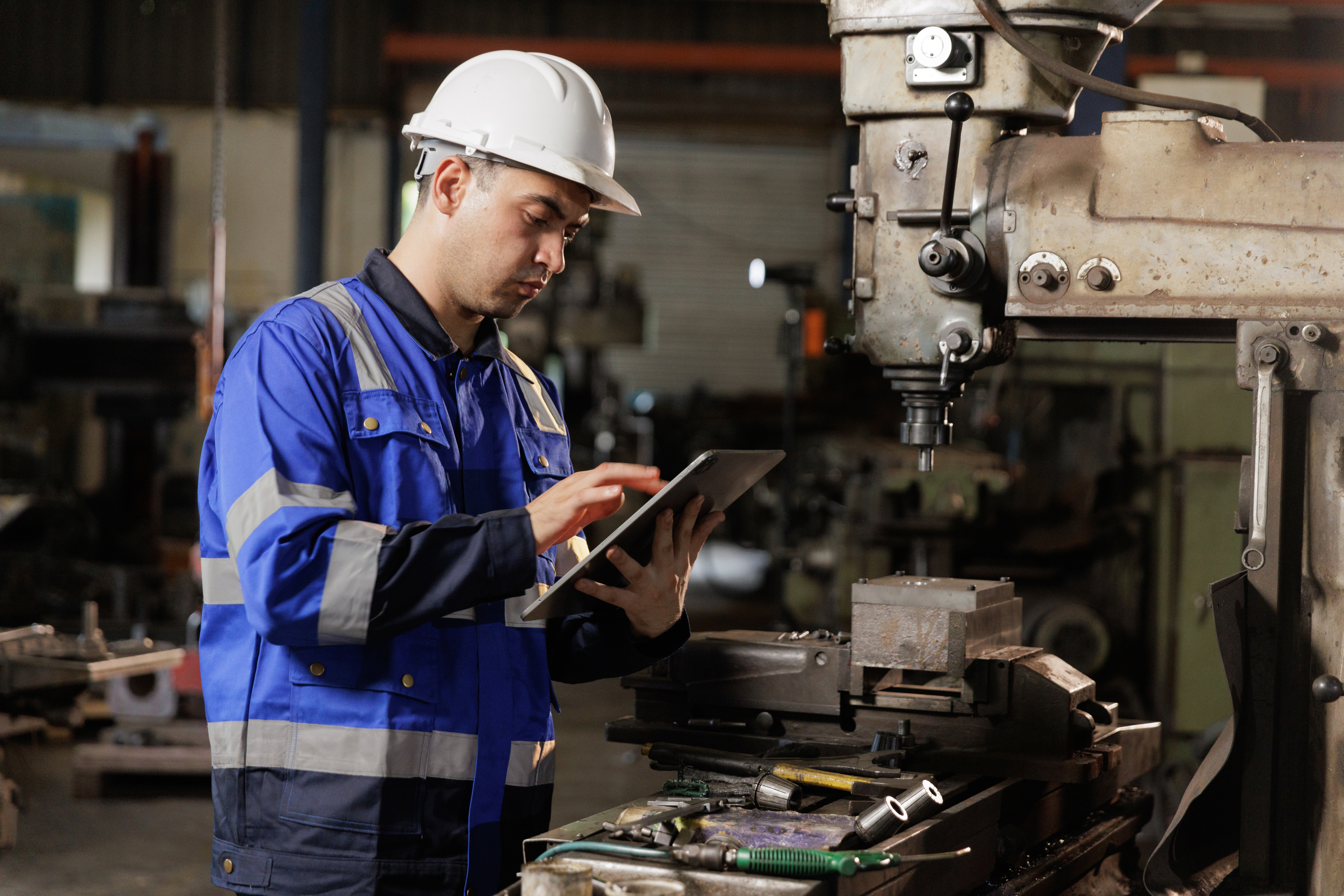 Factory worker in blue safety gear checks tablet near industrial drilling machine