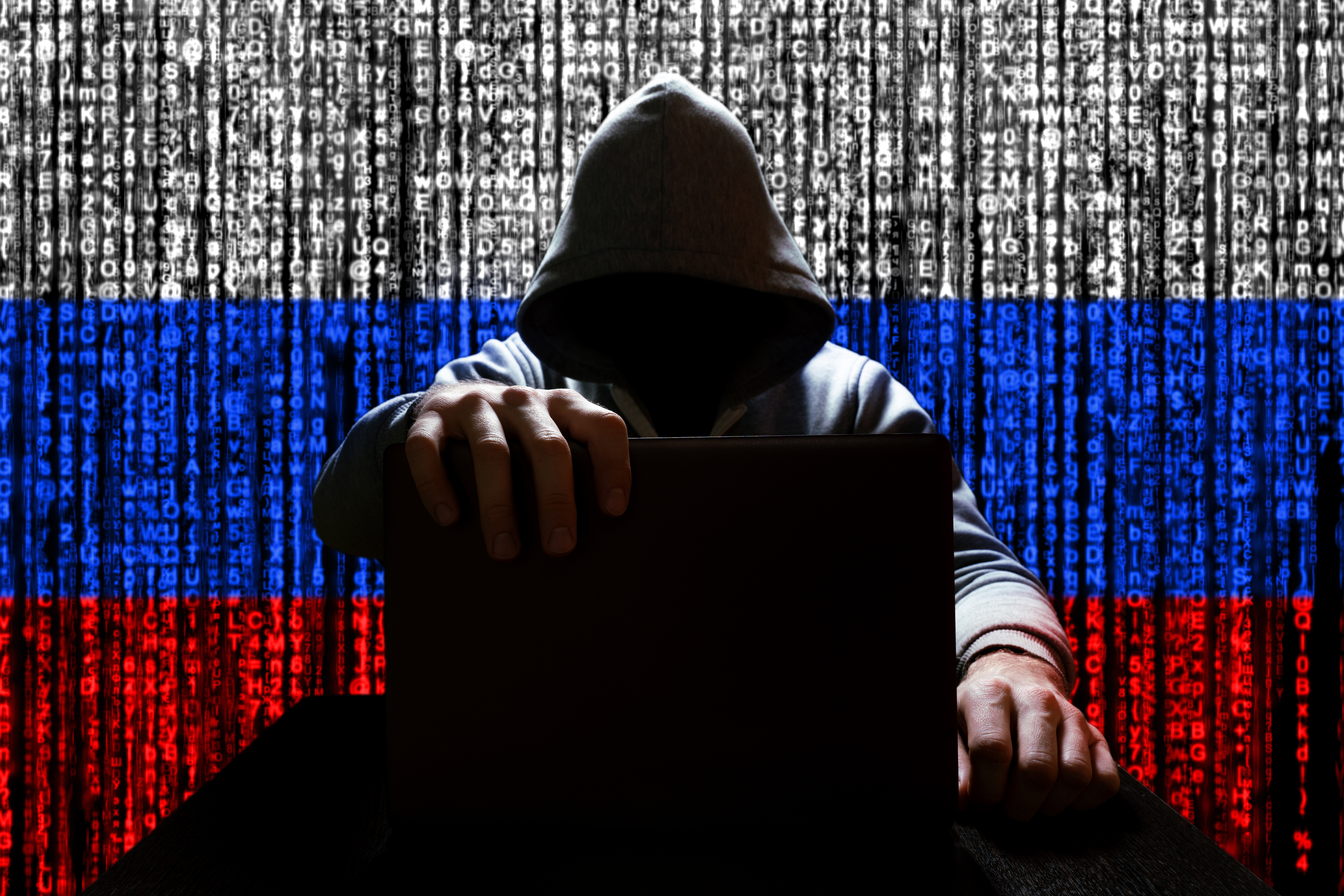 Hooded figure with laptop against Russian flag digital background symbolizing cybersecurity threat