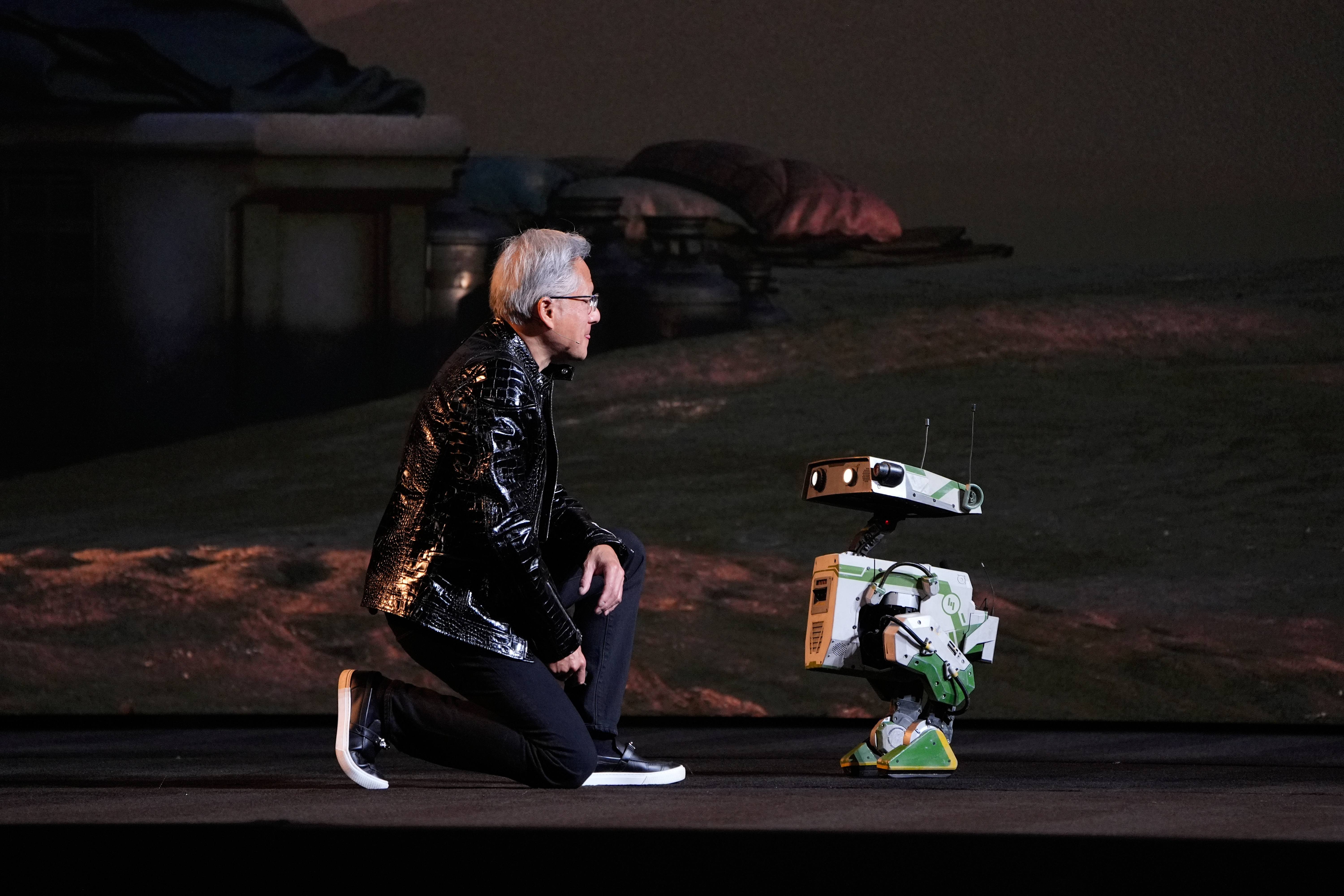 Person in black jacket sitting next to small robotic device on dark stage