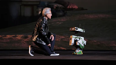 Person in black jacket sitting next to small robotic device on dark stage