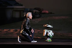 Person in black jacket sitting next to small robotic device on dark stage