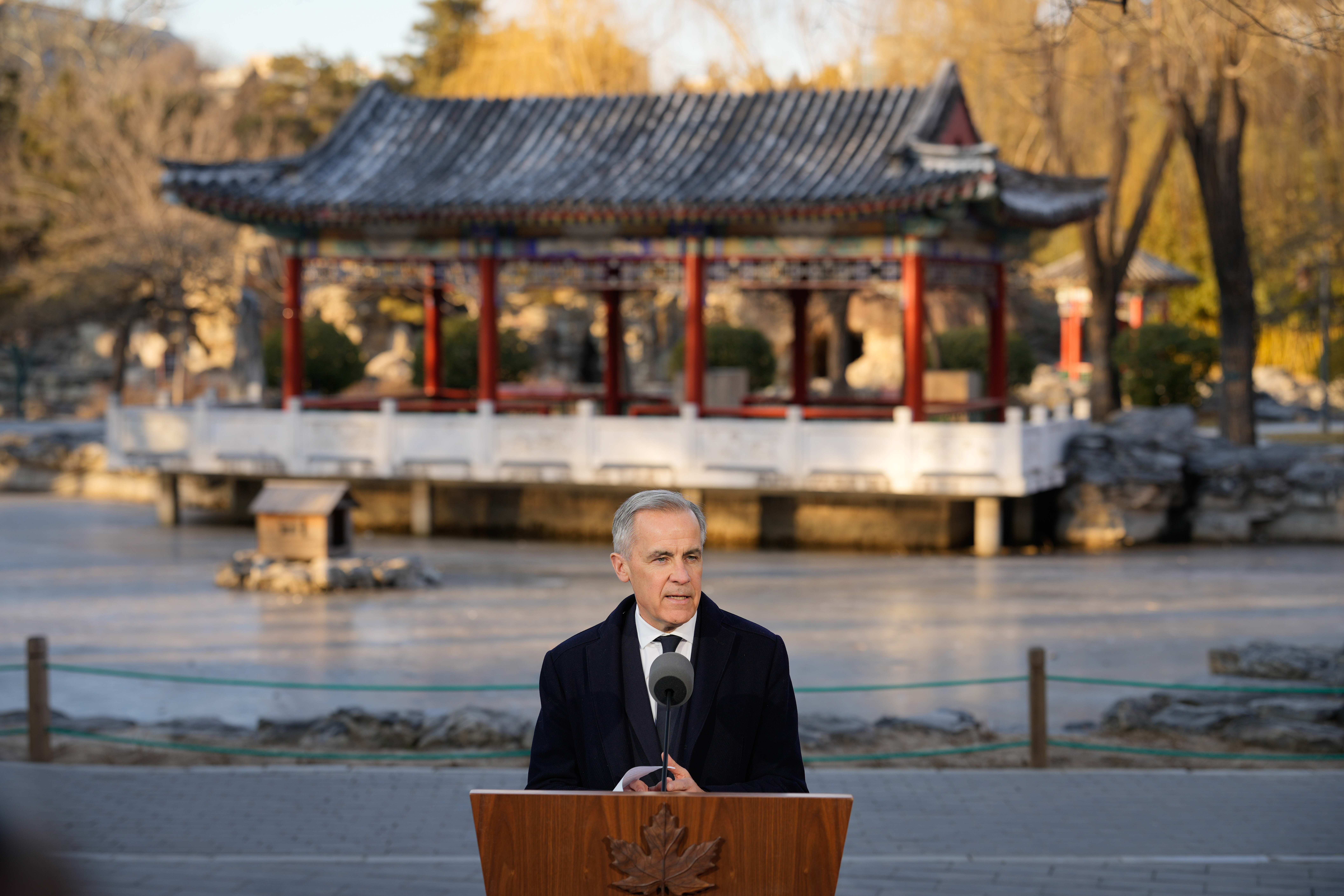 Government official speaking at podium near traditional Chinese pavilion in autumn