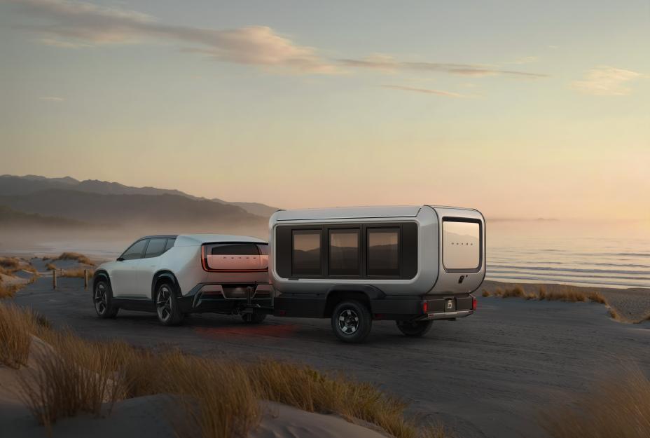 White electric vehicle towing a compact camper trailer on a misty coastal road at sunset