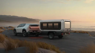 White electric vehicle towing a compact camper trailer on a misty coastal road at sunset