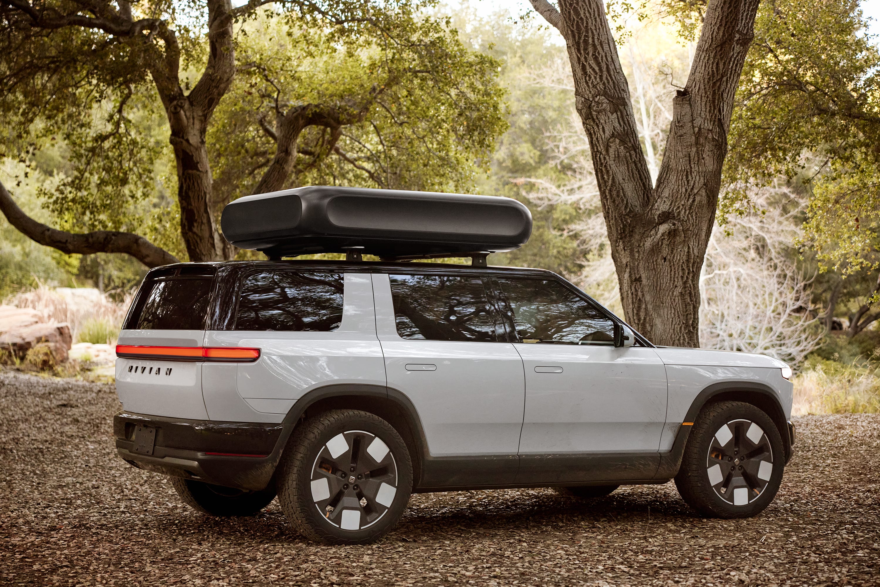 Silver Rivian electric SUV parked on gravel under trees with roof cargo box