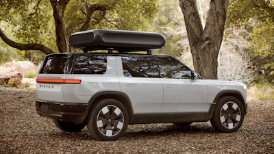 Silver Rivian electric SUV parked on gravel under trees with roof cargo box