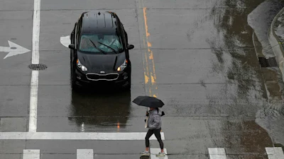 Black Kia car stopped at crosswalk during rainy day, pedestrian with umbrella crossing street