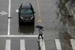 Black Kia car stopped at crosswalk during rainy day, pedestrian with umbrella crossing street