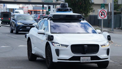 White Waymo autonomous vehicle with blue roof sensors driving on urban street among traffic