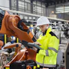 Industrial worker in high-visibility jacket operating orange robotic arm in manufacturing facility