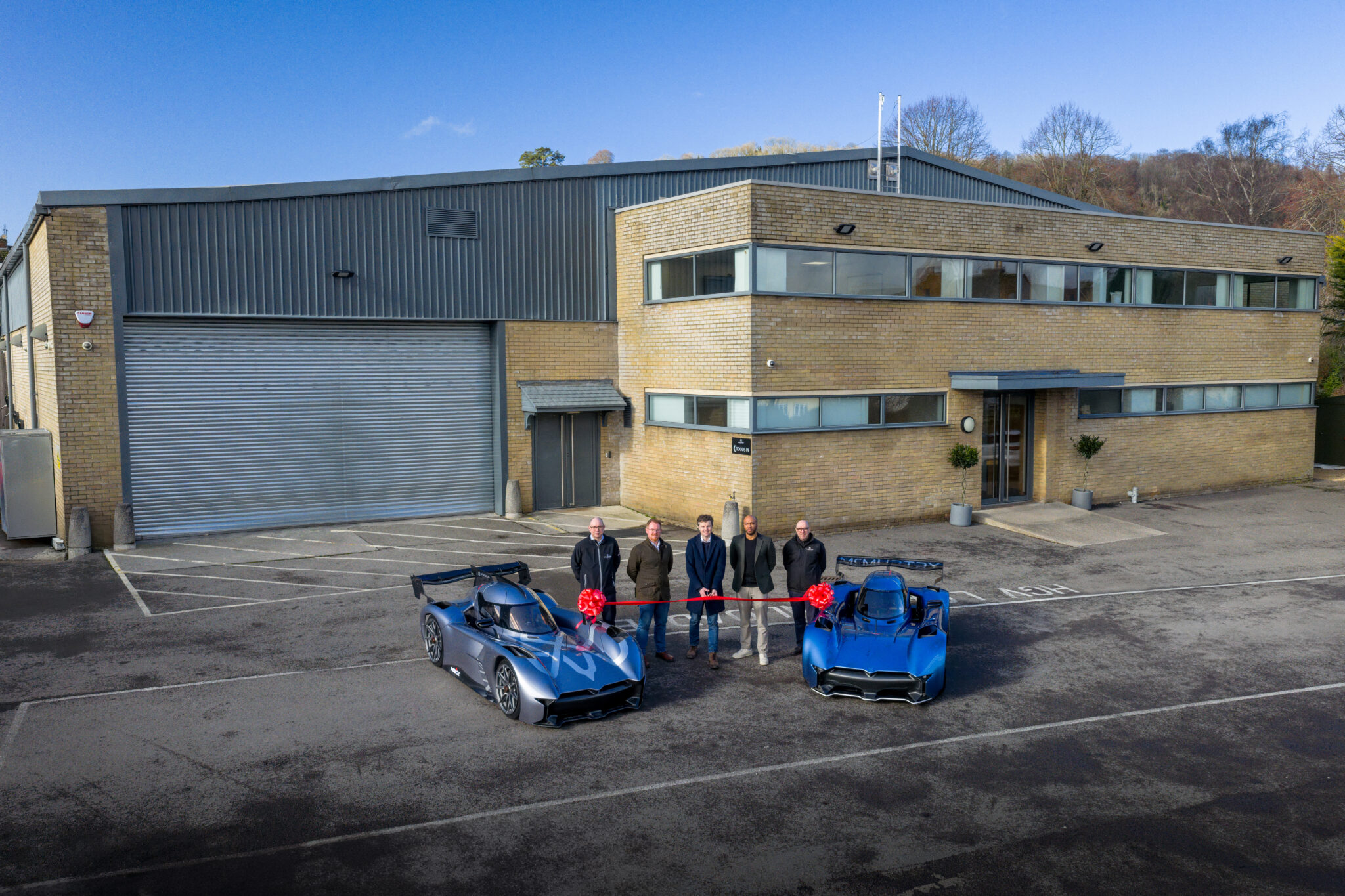 Five people stand between two blue sports cars in front of a modern industrial building with brick and metal cladding.