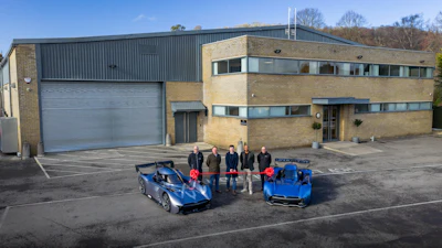 Five people stand between two blue sports cars in front of a modern industrial building with brick and metal cladding.