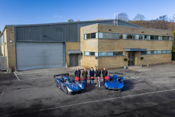 Five people stand between two blue sports cars in front of a modern industrial building with brick and metal cladding.