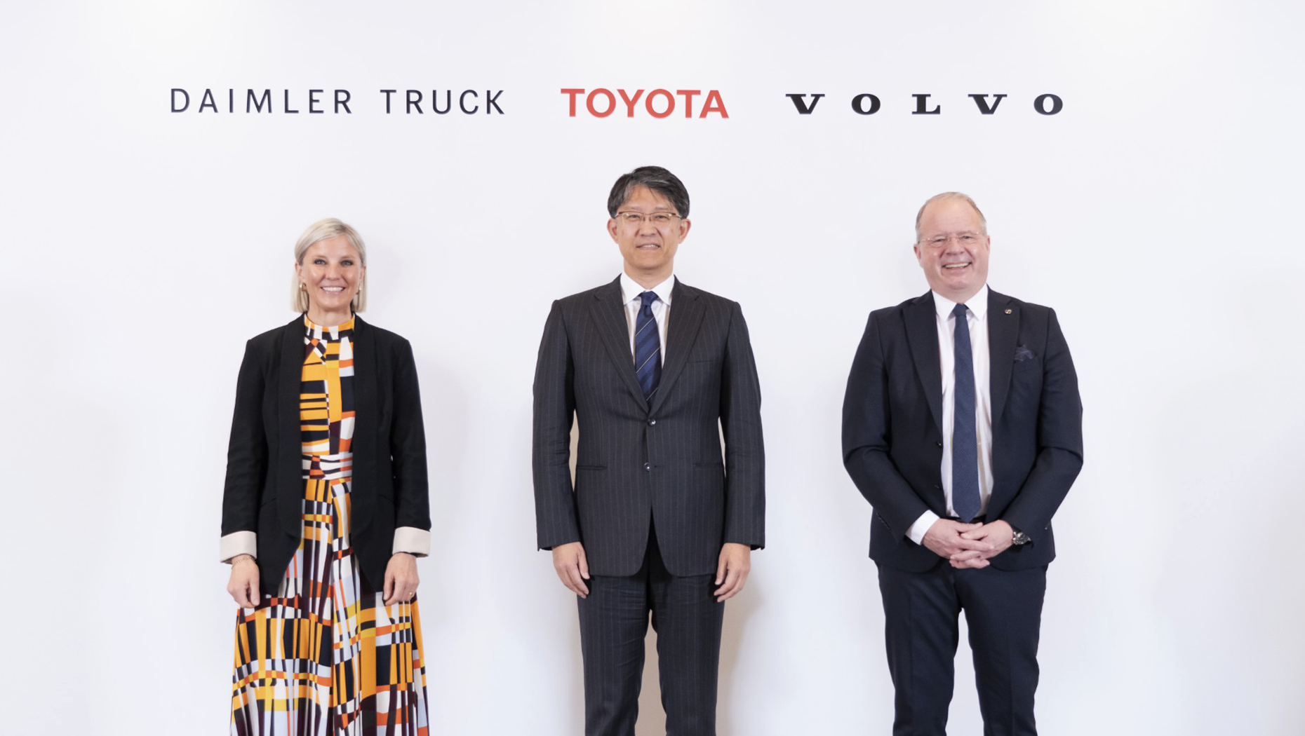 Three business executives pose together at a corporate event with Daimler Truck, Toyota, and Volvo logos displayed behind them