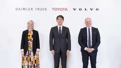 Three business executives pose together at a corporate event with Daimler Truck, Toyota, and Volvo logos displayed behind them