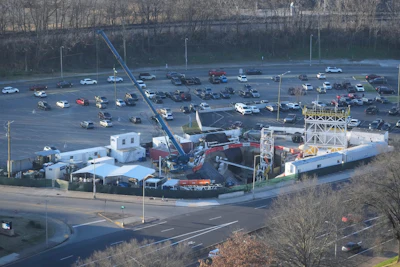 Aerial view of industrial facility with large parking lot, blue crane, and warehouse buildings surrounded by bare trees