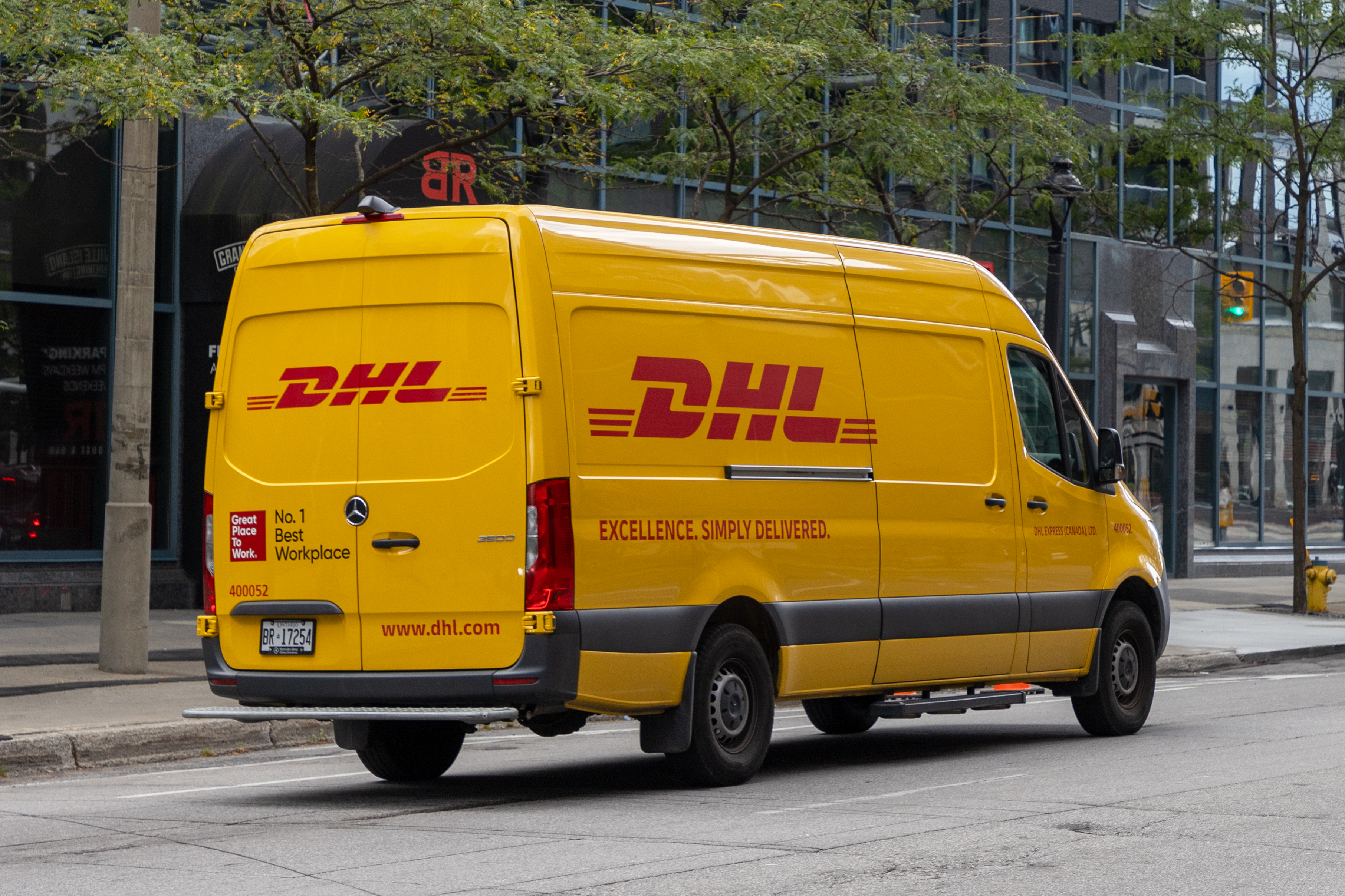 Yellow DHL delivery van parked on urban street with buildings and trees in background