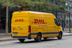 Yellow DHL delivery van parked on urban street with buildings and trees in background