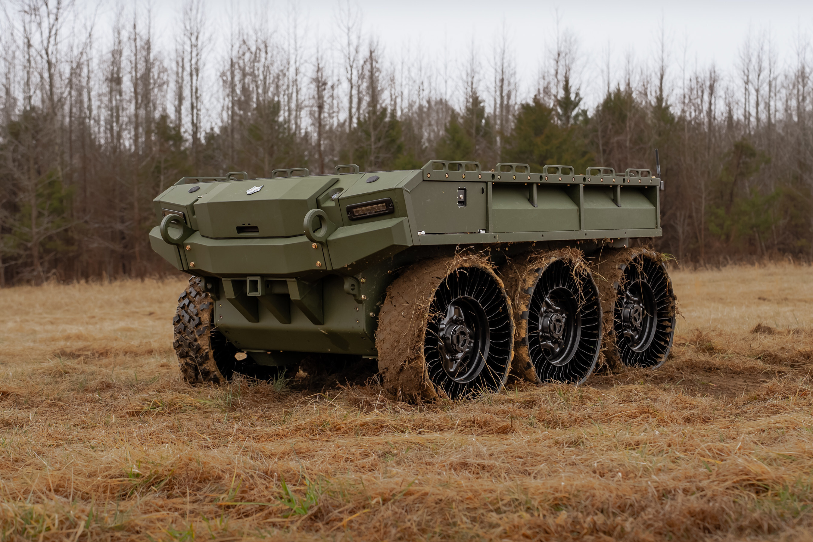 Olive green military six-wheel all-terrain truck with cargo bed parked on dirt terrain with bare trees in background
