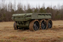 Olive green military six-wheel all-terrain truck with cargo bed parked on dirt terrain with bare trees in background