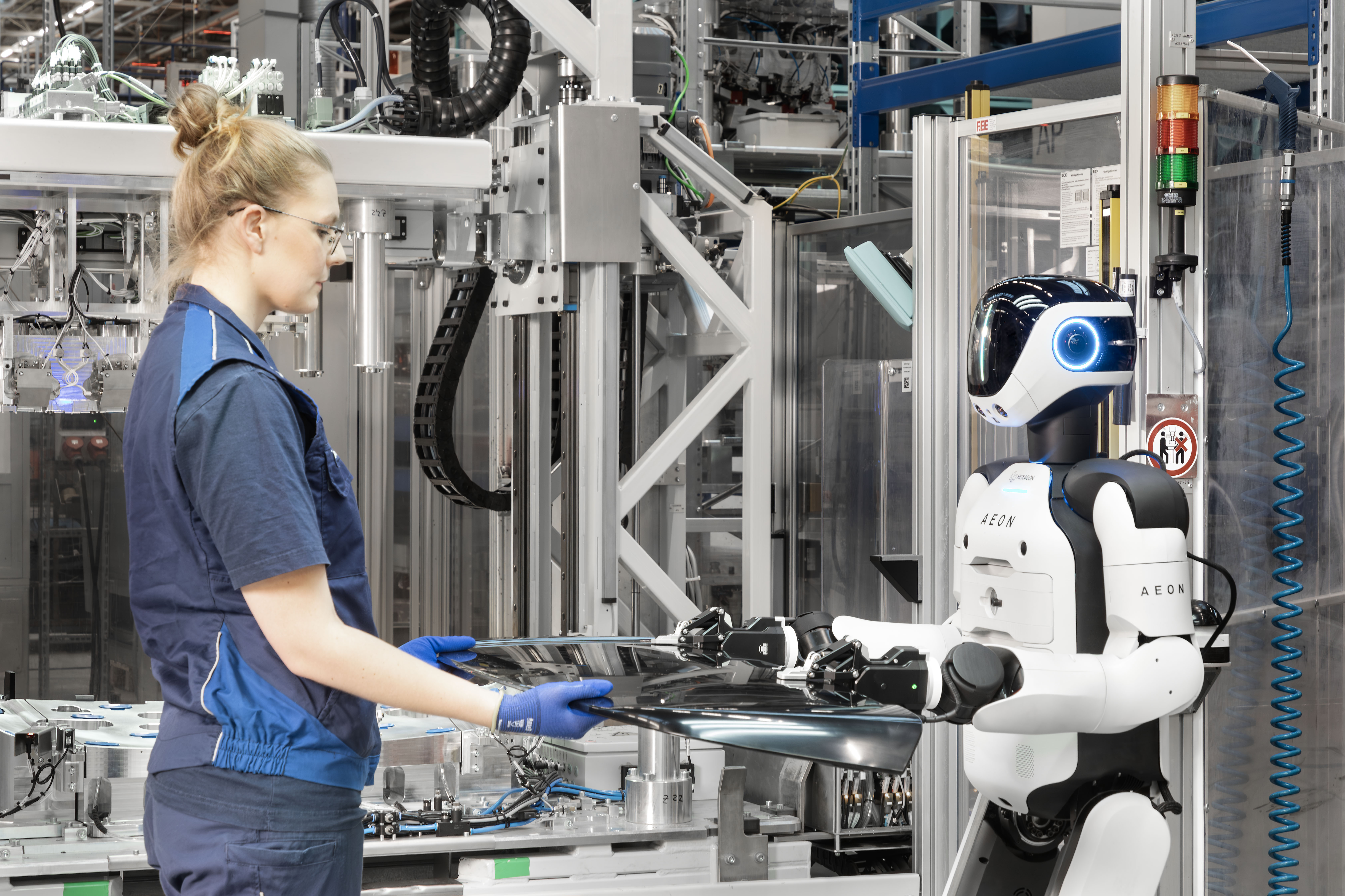 Factory worker collaborating with white and blue AEON humanoid robot at industrial assembly workstation