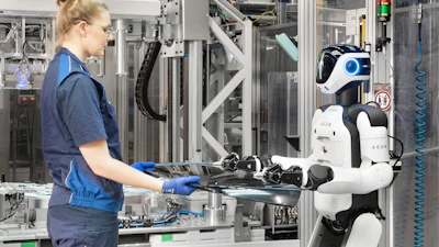 Factory worker collaborating with white and blue AEON humanoid robot at industrial assembly workstation