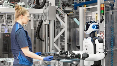 Female technician in blue uniform hands component to white and black humanoid robot in industrial manufacturing facility