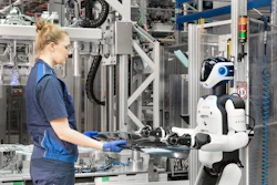 Female technician in blue uniform hands component to white and black humanoid robot in industrial manufacturing facility