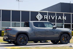 Gray Rivian R1T electric pickup truck parked in front of Rivian facility with company logo signage
