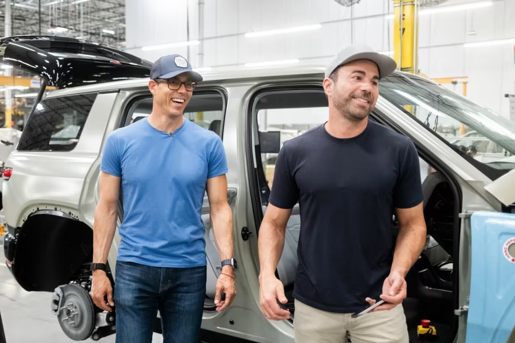 Two men in casual attire stand beside an open vehicle door in an industrial warehouse facility