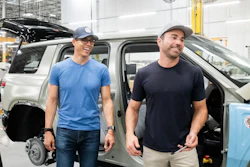 Two men in casual attire stand beside an open vehicle door in an industrial warehouse facility