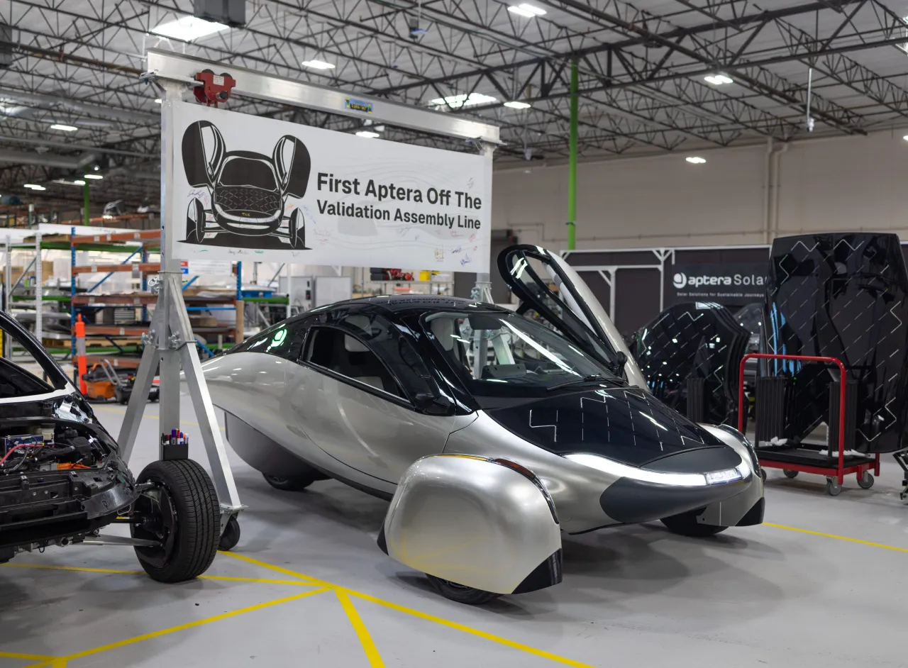 Aptera three-wheeled electric vehicle on display at manufacturing facility with validation assembly line banner