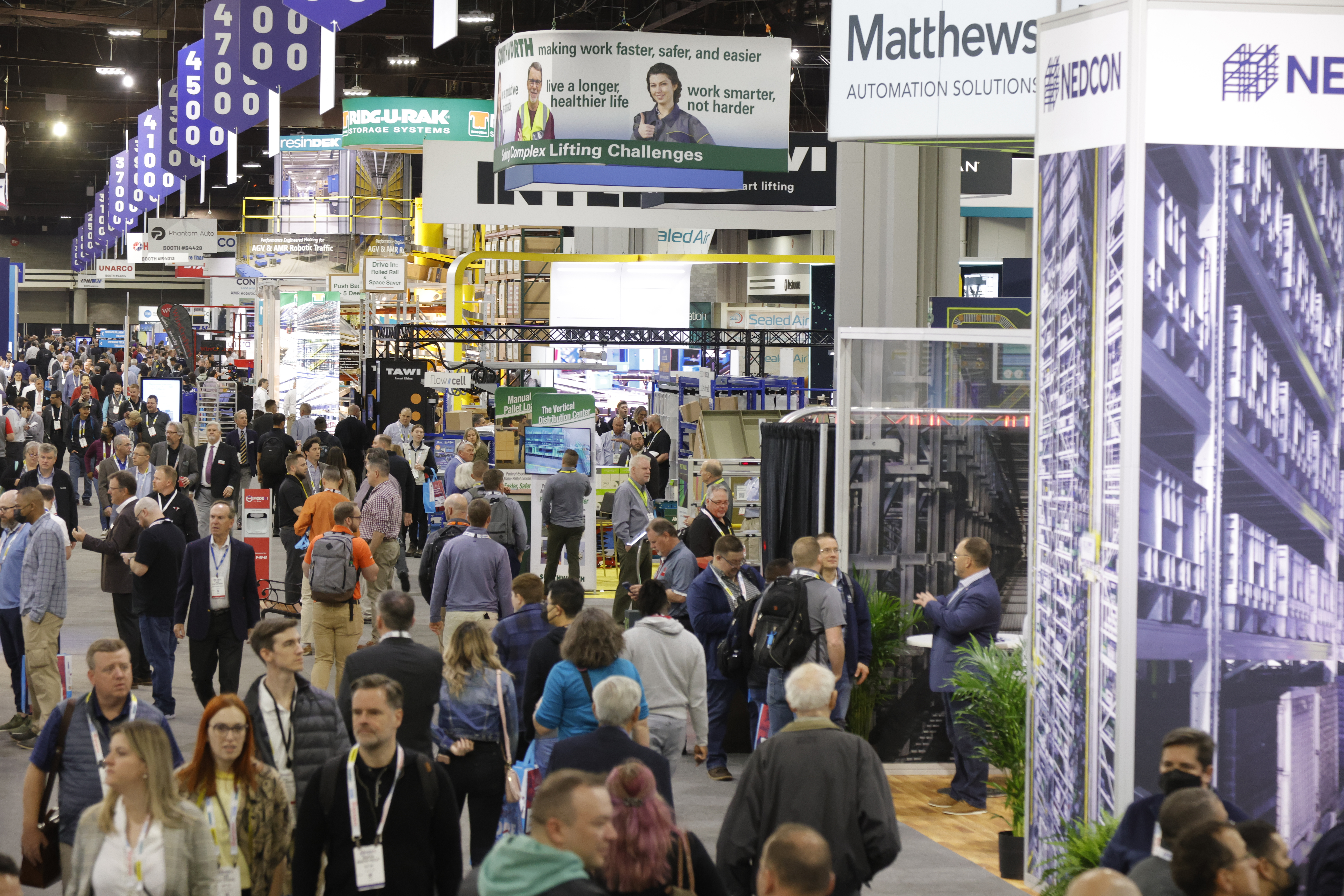 Crowded trade show floor with multiple vendor booths, purple and blue signage, and attendees networking among exhibition displays.