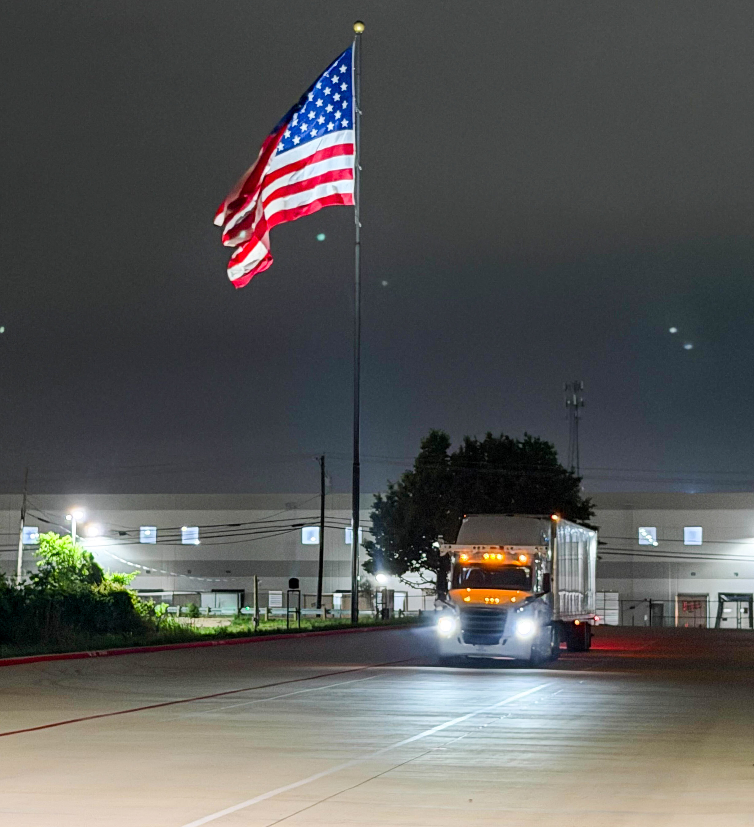 American flag illuminated at night on flagpole above industrial facility with truck and buildings
