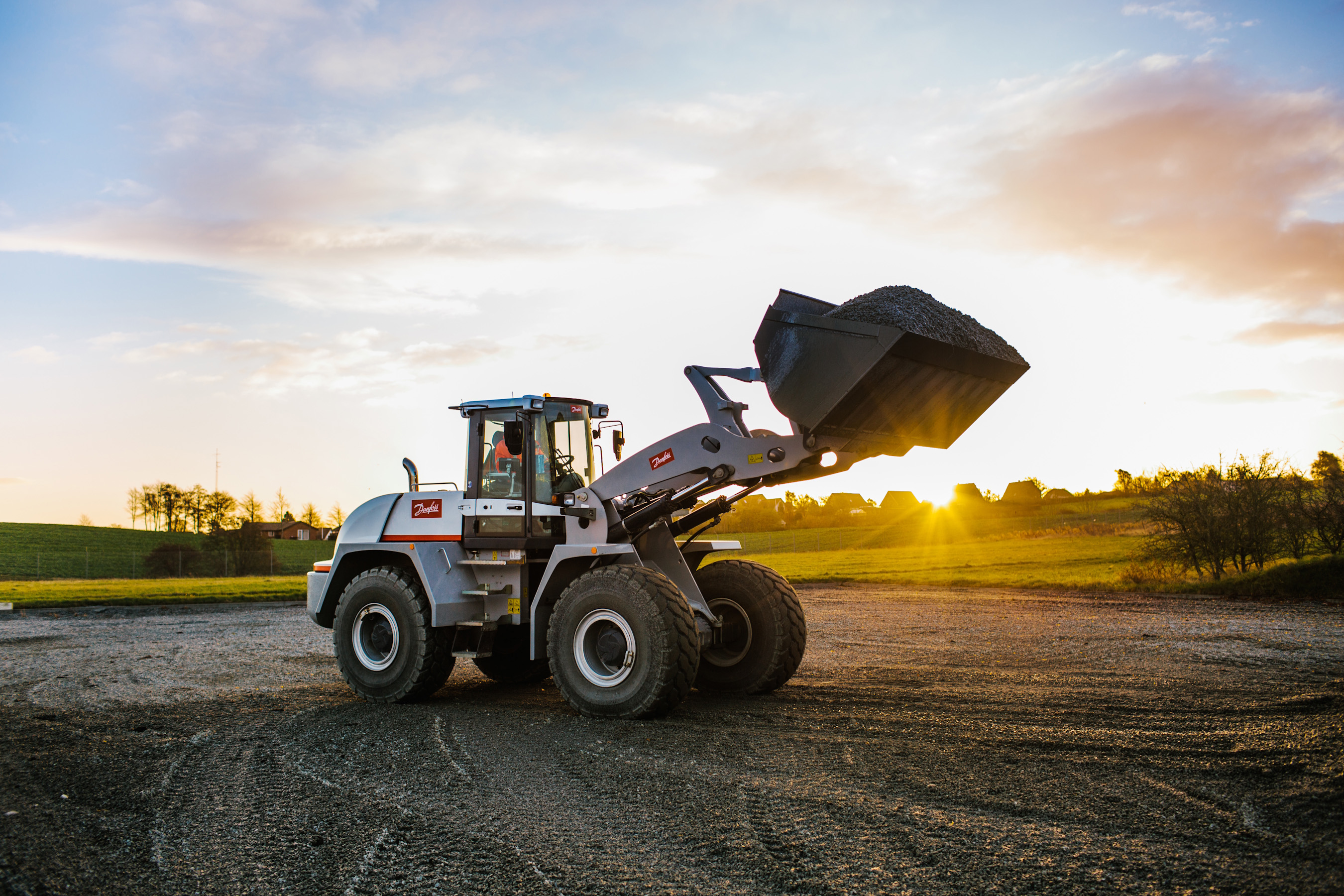 Wheel loader with raised bucket on rural road at sunset with green fields in background