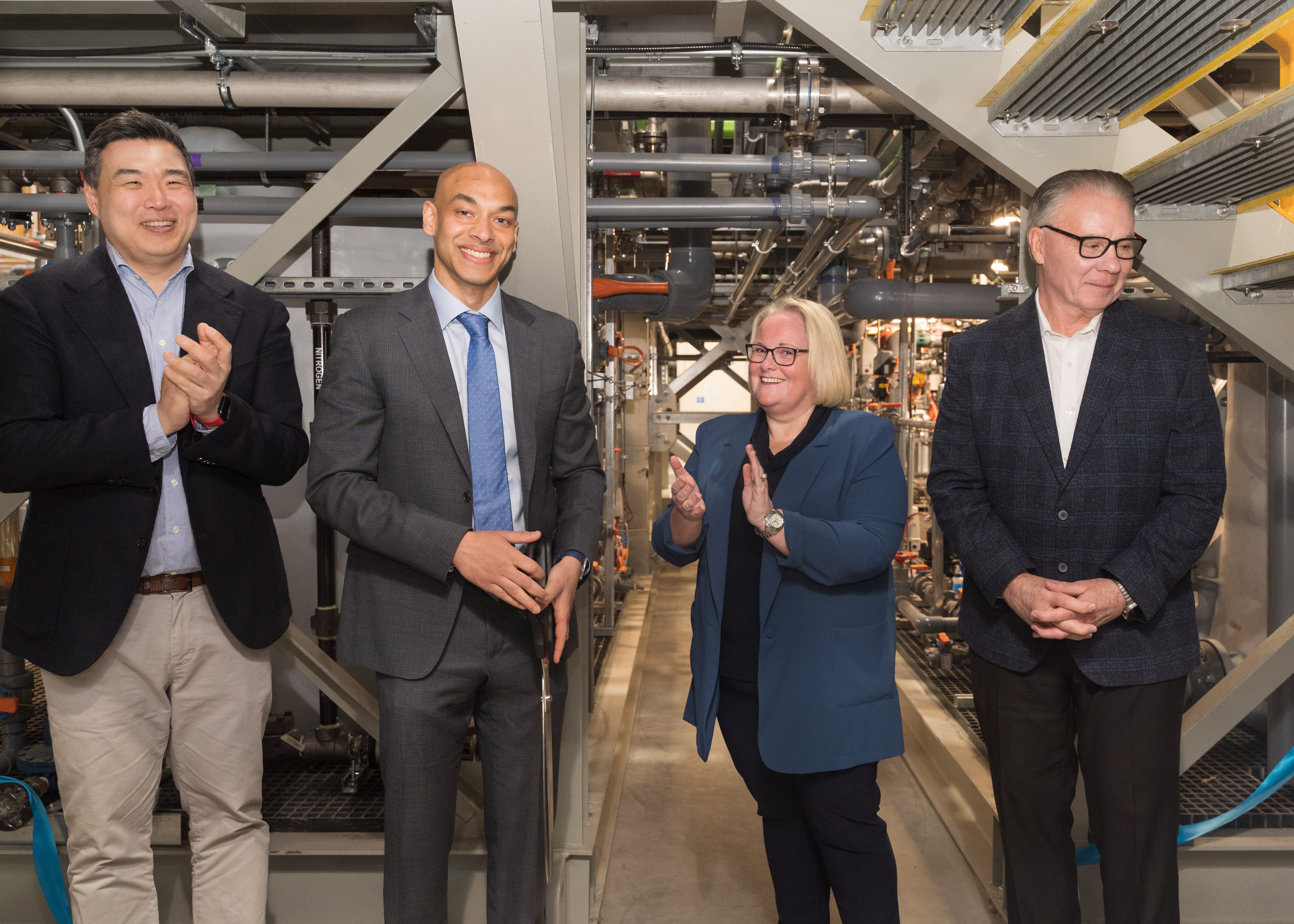 Four business professionals in formal attire standing together in an industrial facility with machinery and equipment visible in the background.