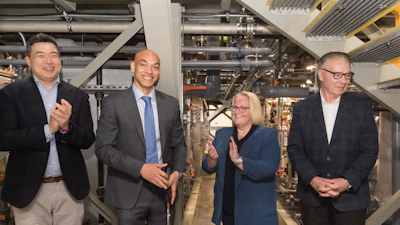 Four business professionals in formal attire standing together in an industrial facility with machinery and equipment visible in the background.